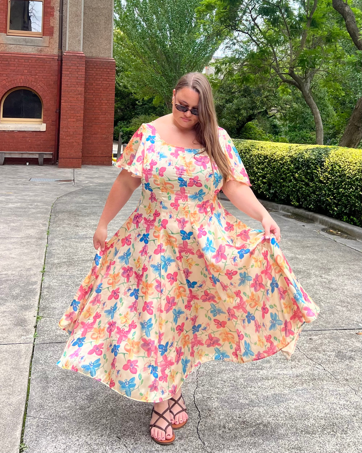 Model wearing Lucille Dress in Satin Floral print, front view twirling skirt, flutter sleeve midi dress with pink, blue and orange florals, sunglasses, against brick building