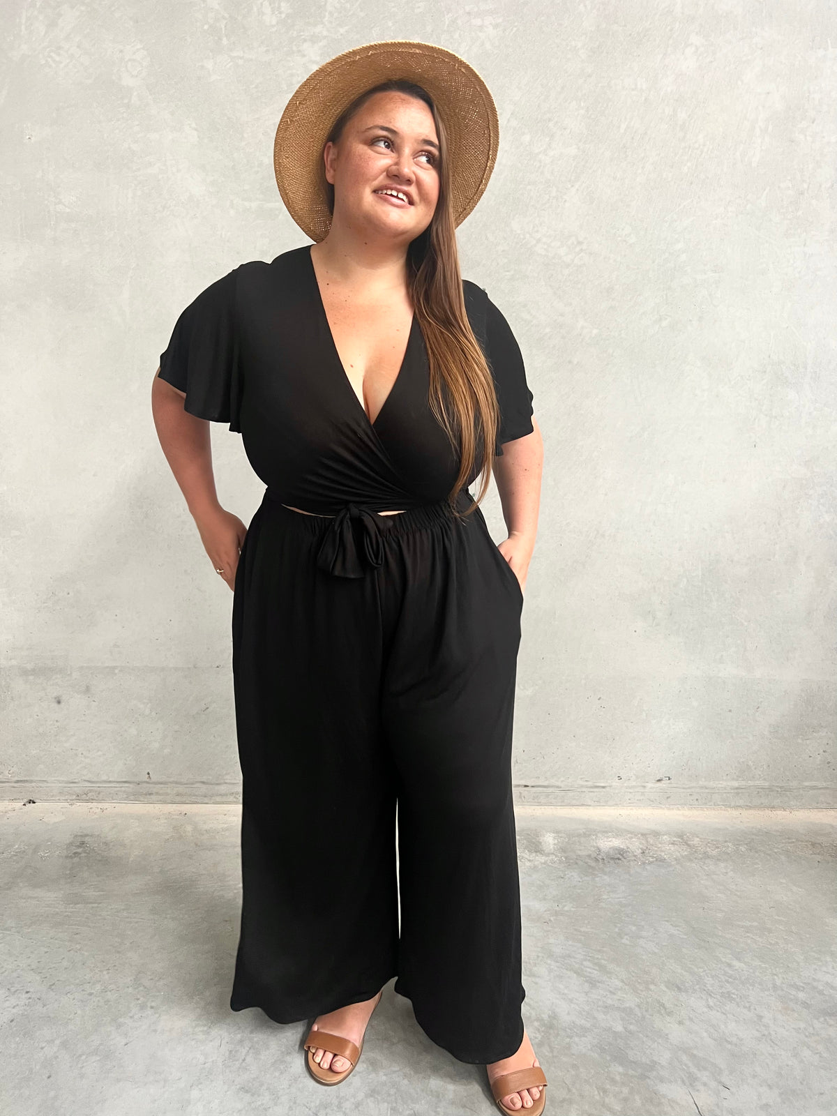 Model in black Vivienne Wrap Top with matching wide-leg pants and straw hat, concrete wall background