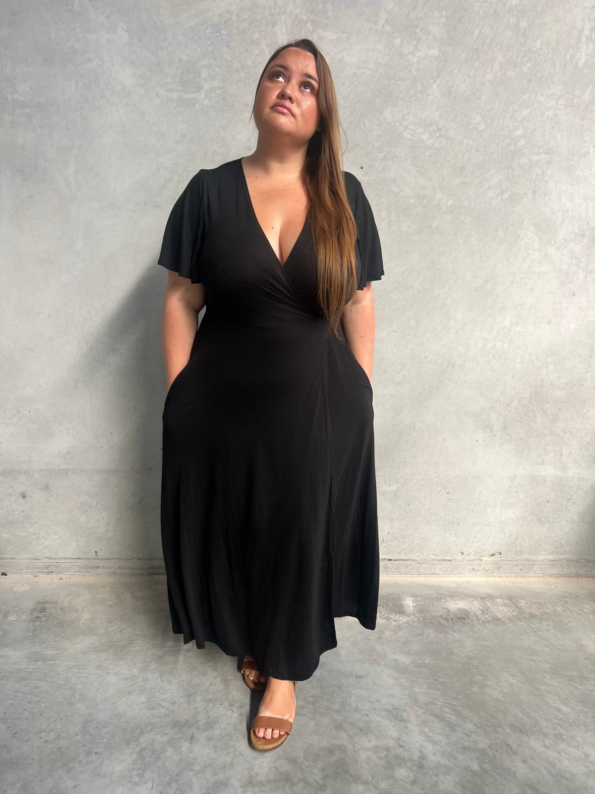 Model in black Belle Wrap Dress with flutter sleeves and V-neckline, concrete wall background