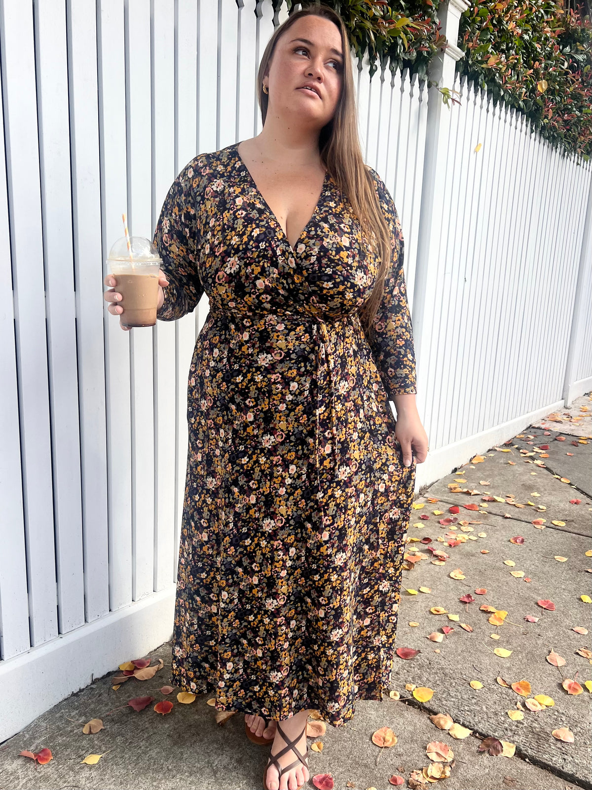 Model in navy floral long-sleeve Alexia Wrap Dress holding an iced coffee, white picket fence background