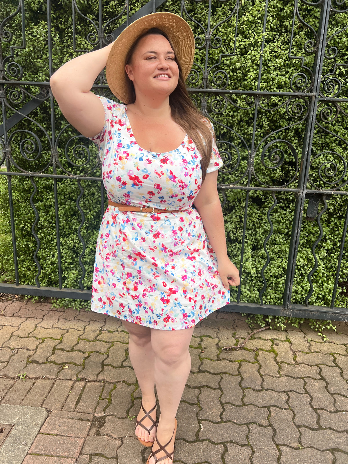 Model in floral Arianna Dress with tan belt and straw hat, standing by an iron gate