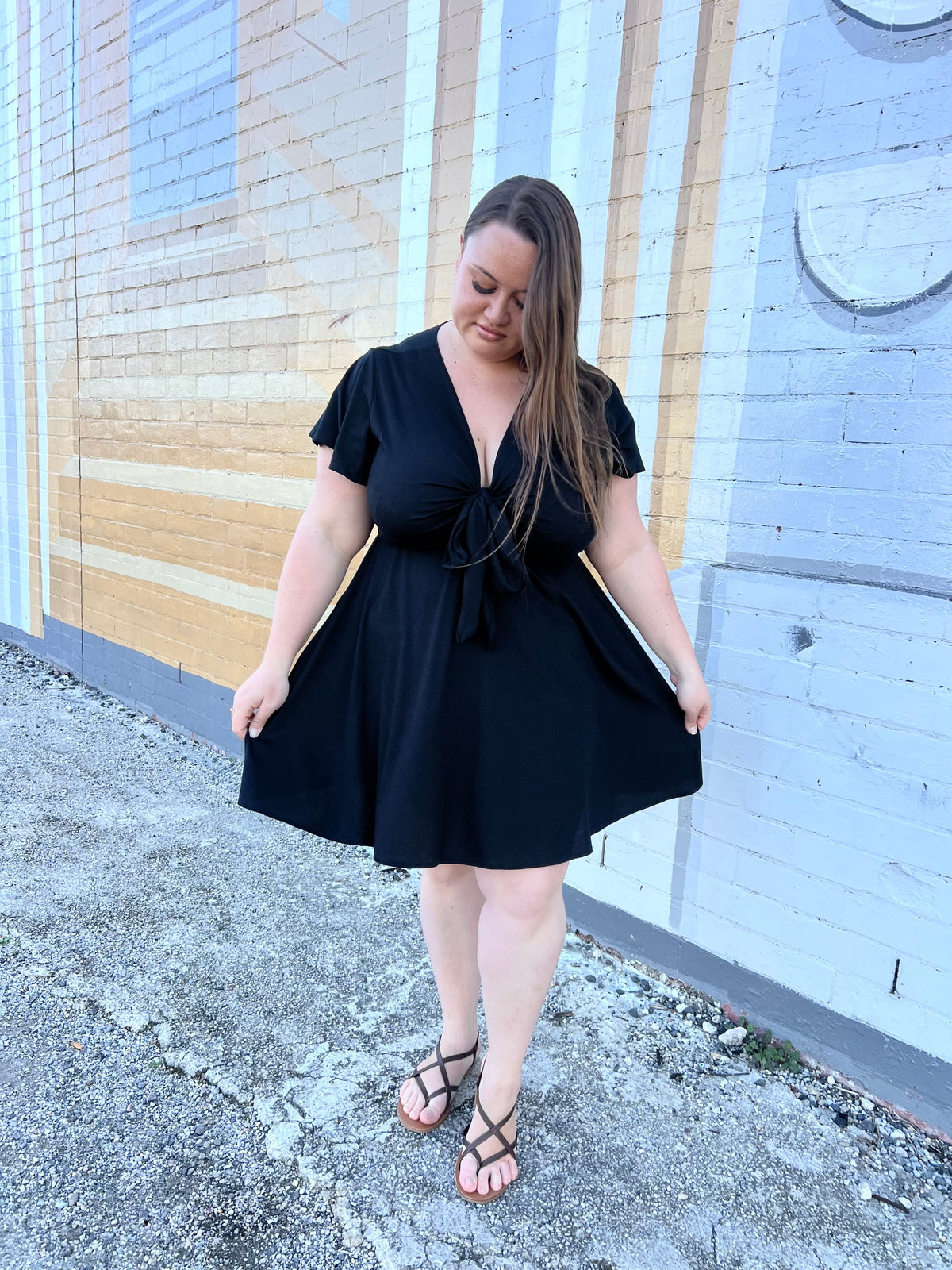 Model wearing Amber Dress in Black, front view looking down, tie-front V-neck skater dress with flutter sleeves against painted brick wall